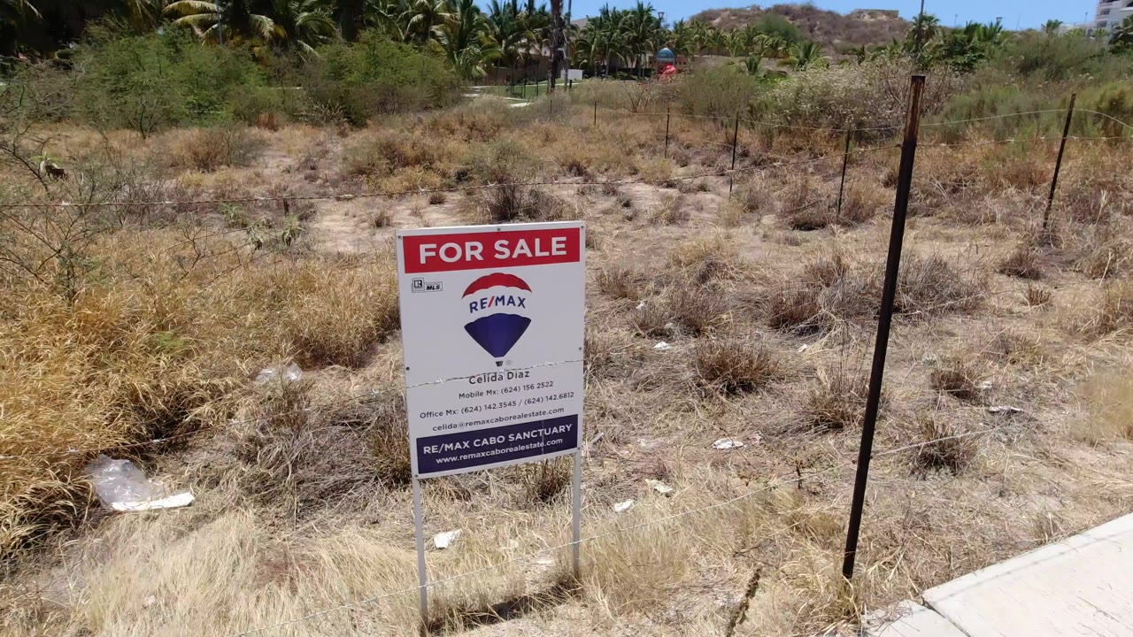 A red and white for-sale sign stands beside a dry fenced lot in San Jose del Cabo, with scrub, palm trees, low hills, and no visible people.