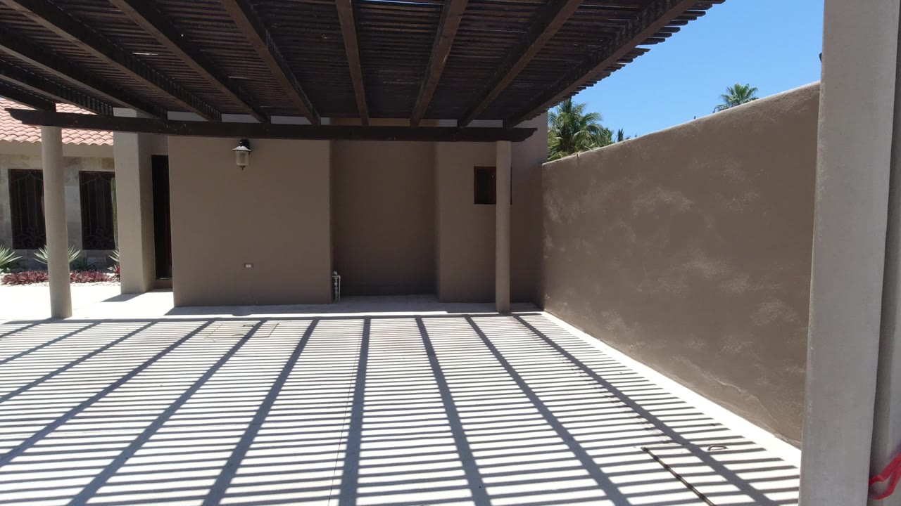 Sunlit stucco courtyard in San Jose del Cabo with a slatted pergola overhead, a blank wall, pale paving, and no visible people.