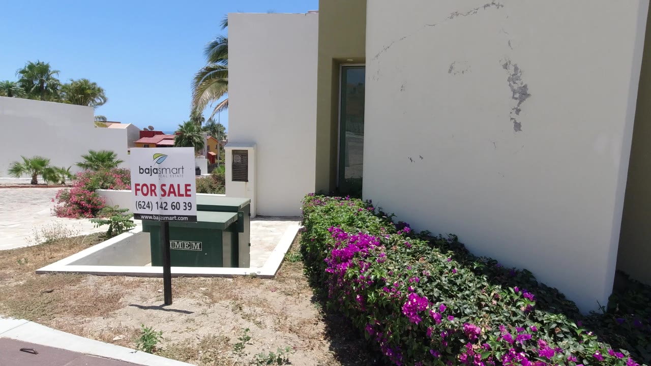 For-sale sign and green dumpster beside a modern white building with flowering shrubs.