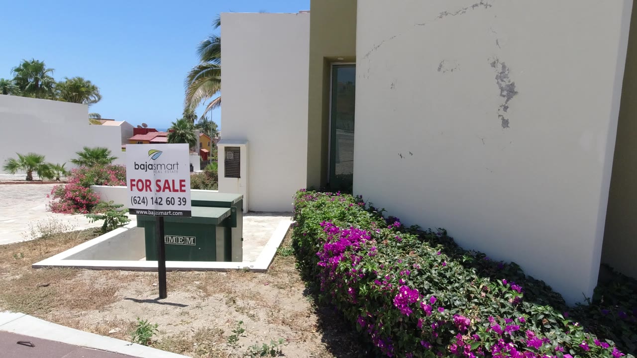The for-sale sign and green dumpster remain in the foreground as the white building wall and flowering hedge fill the San Jose del Cabo frame, with no visible people.