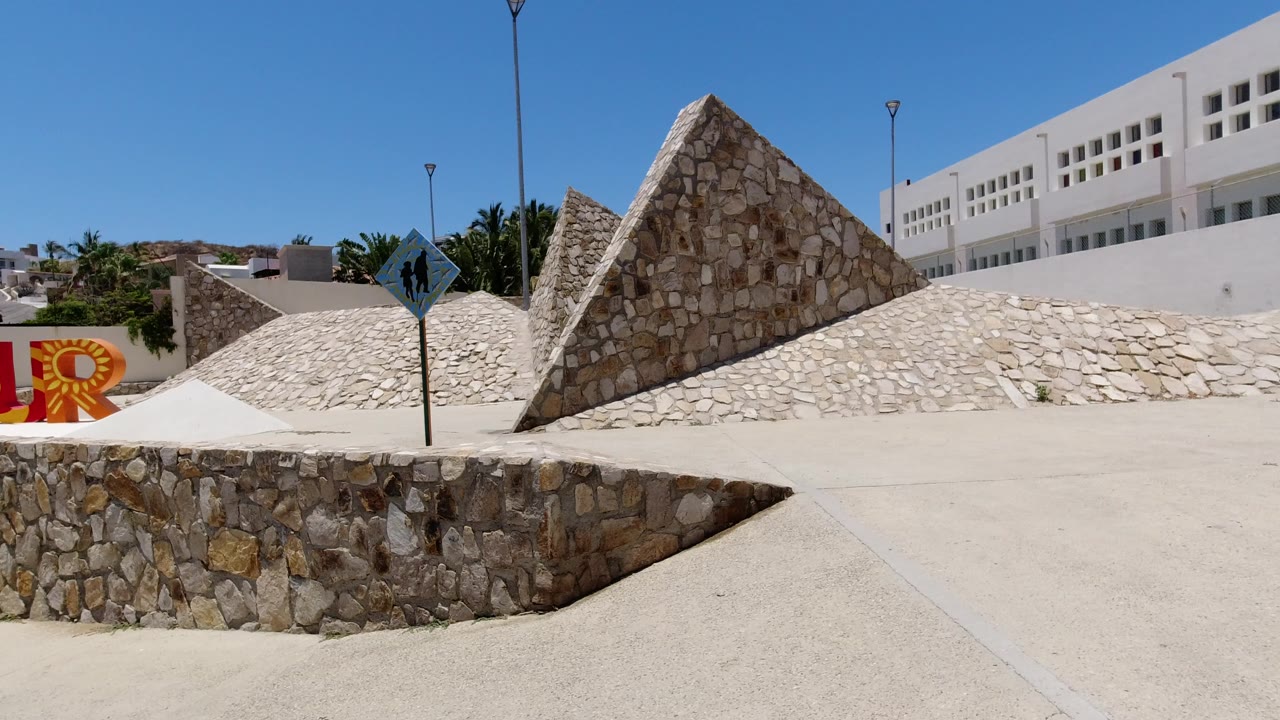 Stone plaza with pyramid-like walls, white building, blue sky, and partial colorful letters.