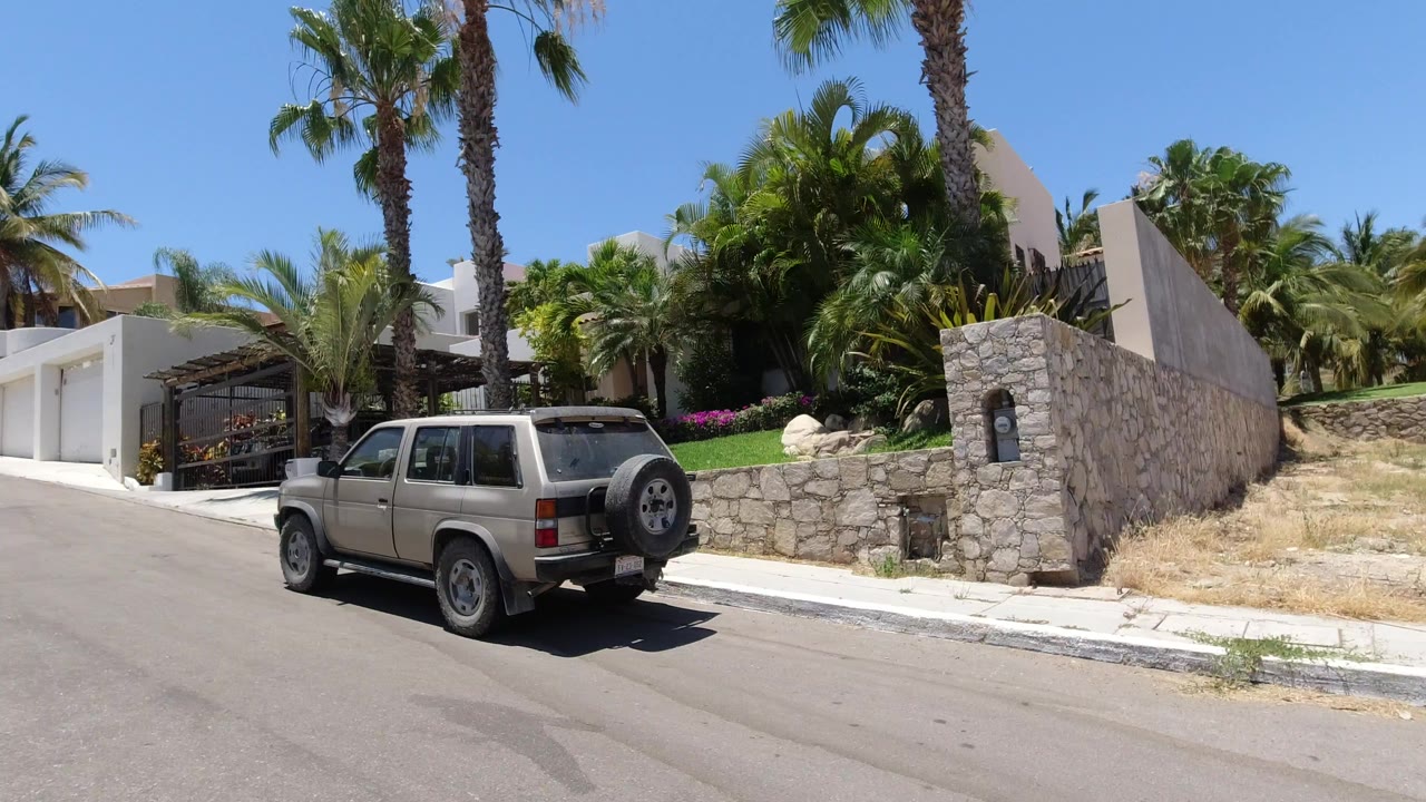 A beige SUV is parked along a sunlit residential street in San Jose del Cabo, Mexico, with a stone wall, white buildings, palm trees, dry grass, and no visible people.