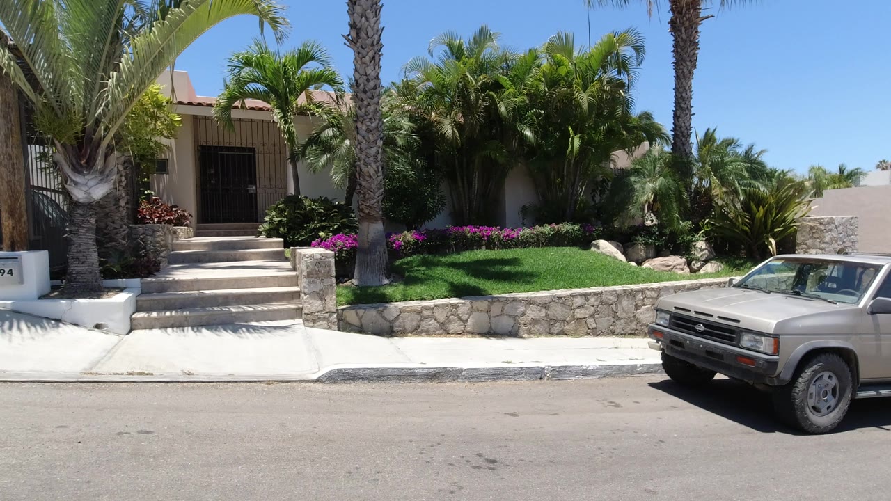 A parked vehicle sits beside the curb in front of a San Jose Del Cabo home with palms, trimmed grass, flowering plants, stonework, and no visible people.