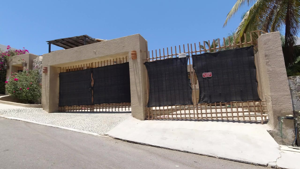 A San Jose del Cabo building entrance with beige walls, black gates, white gravel, flowering shrubs, palm fronds, and no visible people.