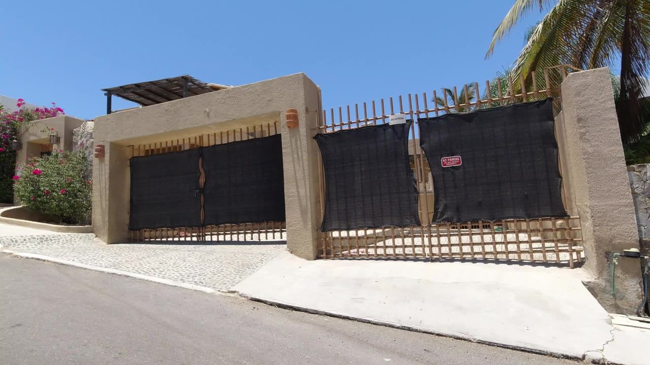 A closer street-facing view of the San Jose del Cabo gate shows black metal panels, beige stucco walls, flowering shrubs, palm leaves, and no visible people.