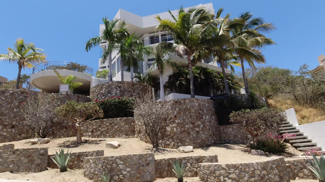 White modern mansion above stone terraces and palm trees under blue sky.