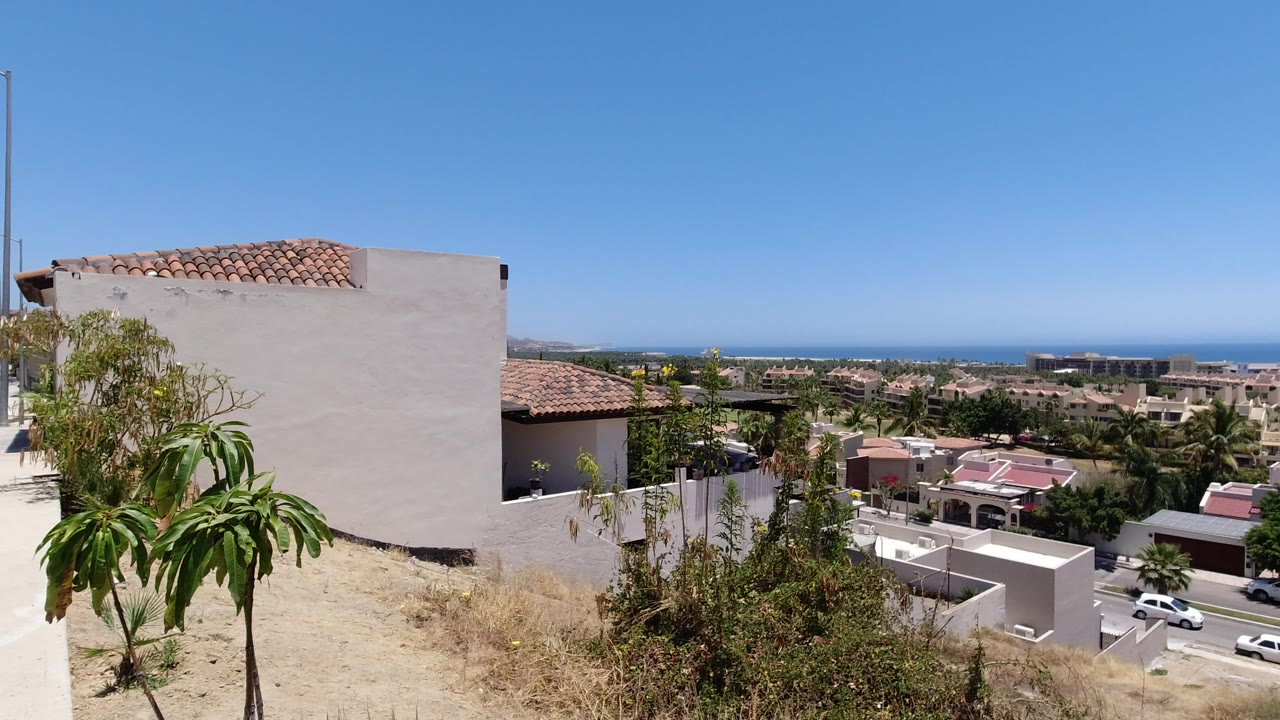 Elevated view of houses, dry hillside plants, and distant sea under a clear sky.
