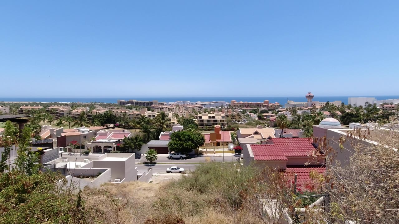 First frame of San Jose del Cabo showing an elevated view over low buildings, palm trees, a road, dry hillside brush, blue ocean, and no visible people.