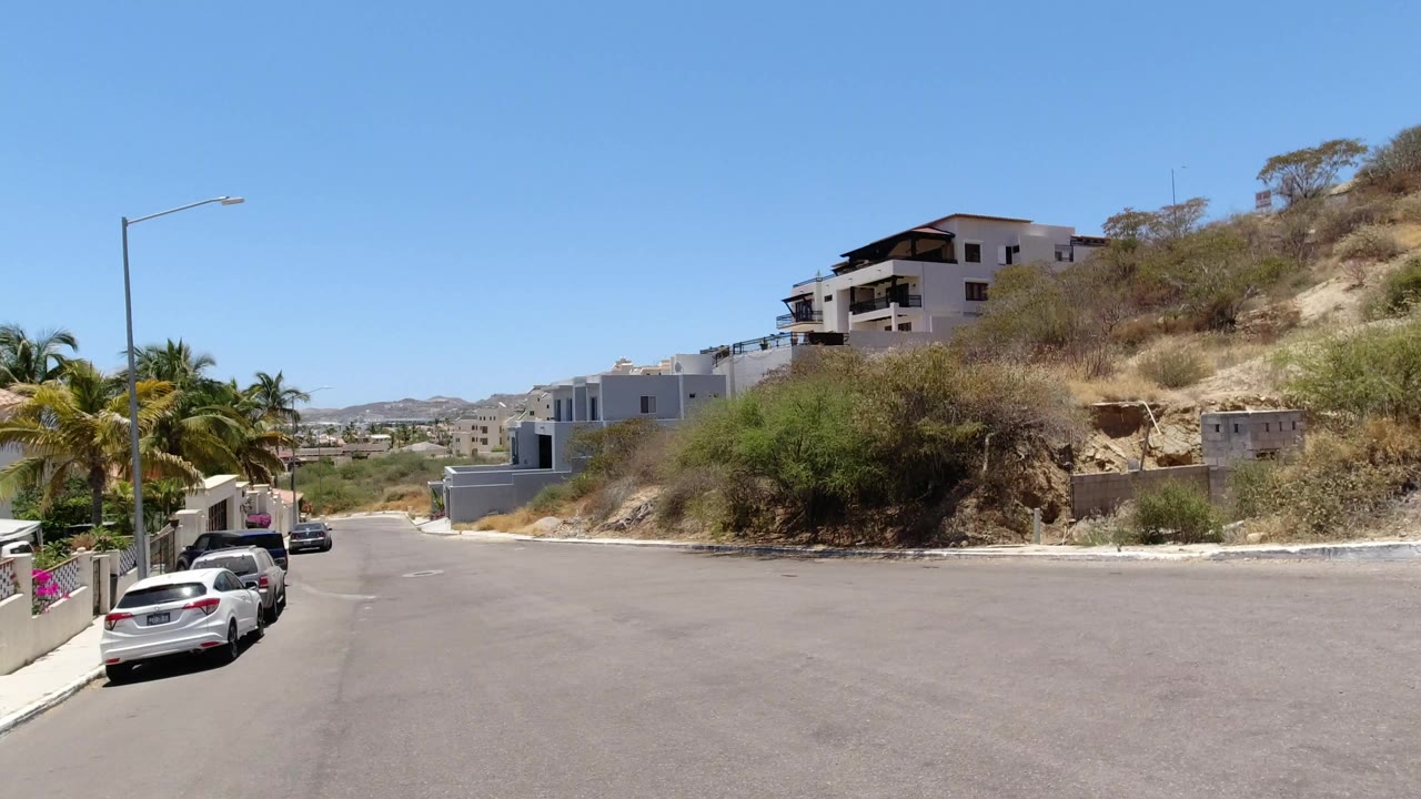 A wide San Jose del Cabo residential street view shows parked cars along the left curb, white homes on the hillside, dry brush, palm trees, and no visible people.