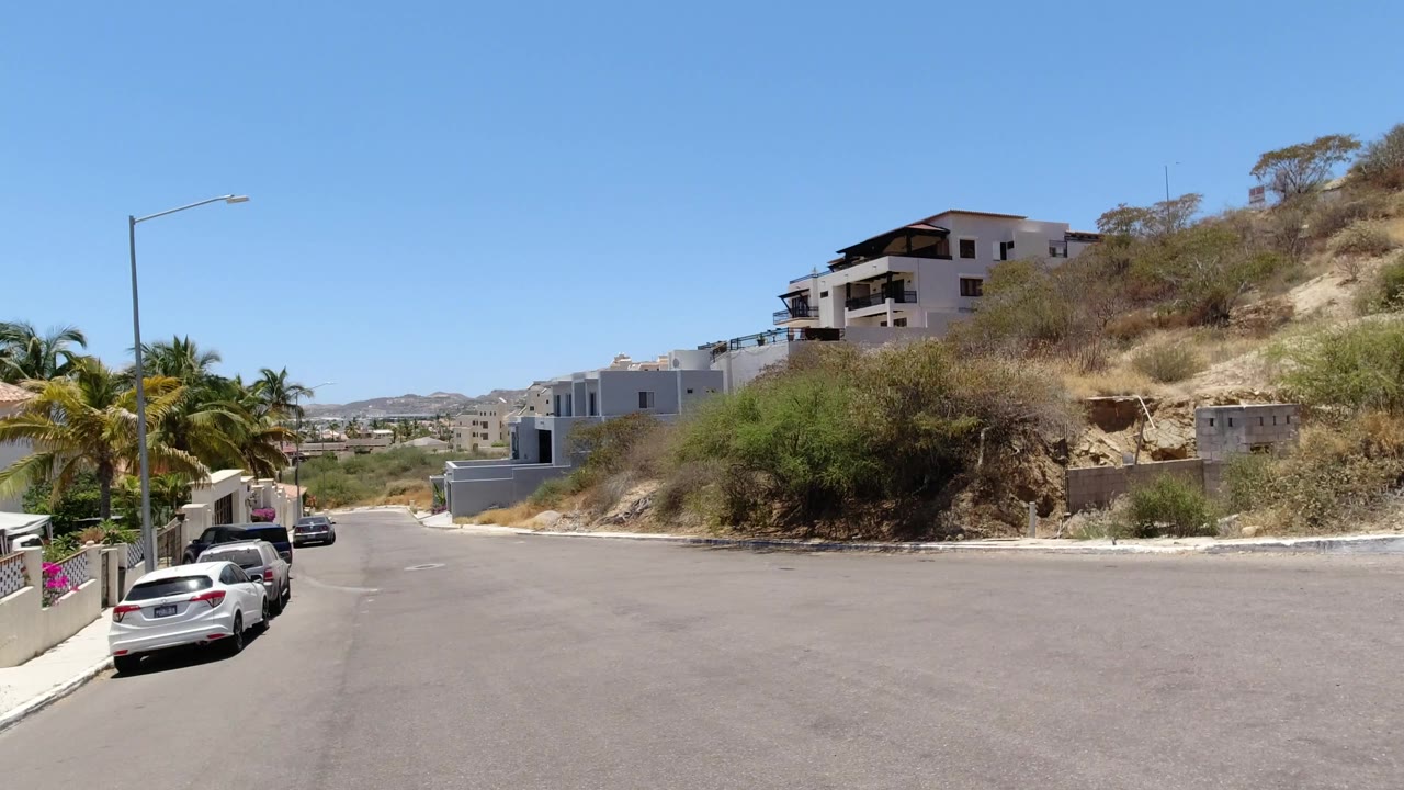 A similar San Jose del Cabo street angle shows a broad asphalt turn, pale hillside homes, dry roadside plants, parked cars on the left, and no visible people.