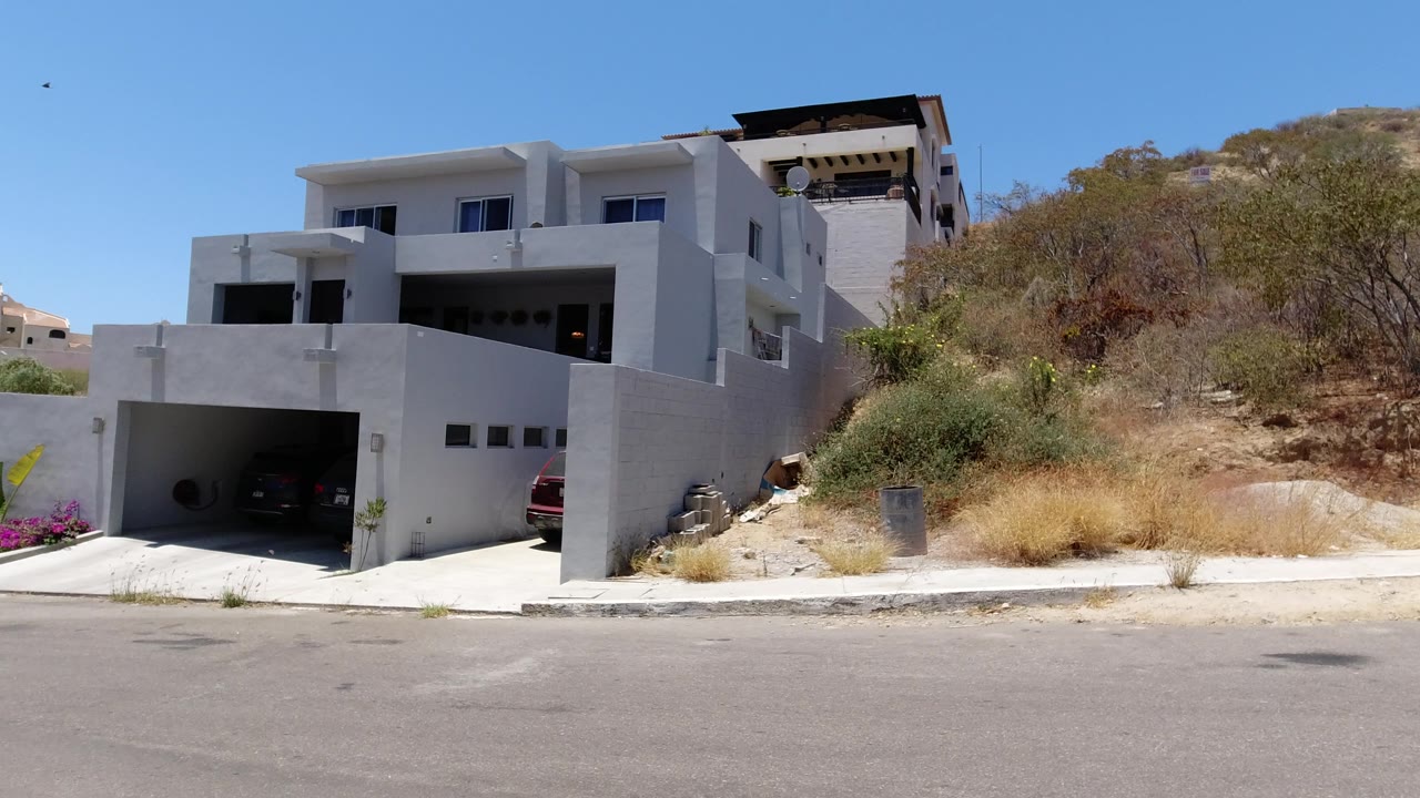 The house remains centered in San Jose del Cabo, with the shaded garage, light concrete driveway, dry brushy slope, and no visible people.