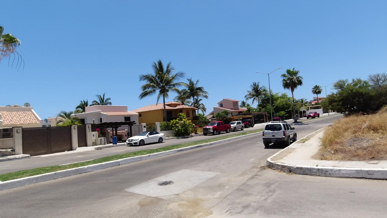 A San Jose Del Cabo street view with a white car near a gated home, red vehicles farther back, palms over low houses, dry roadside grass, and no visible people.