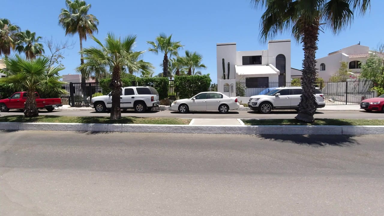 Sunny San Jose del Cabo residential street with palms, gated homes, a white SUV, a white sedan, and a red pickup across the road, with no visible people.