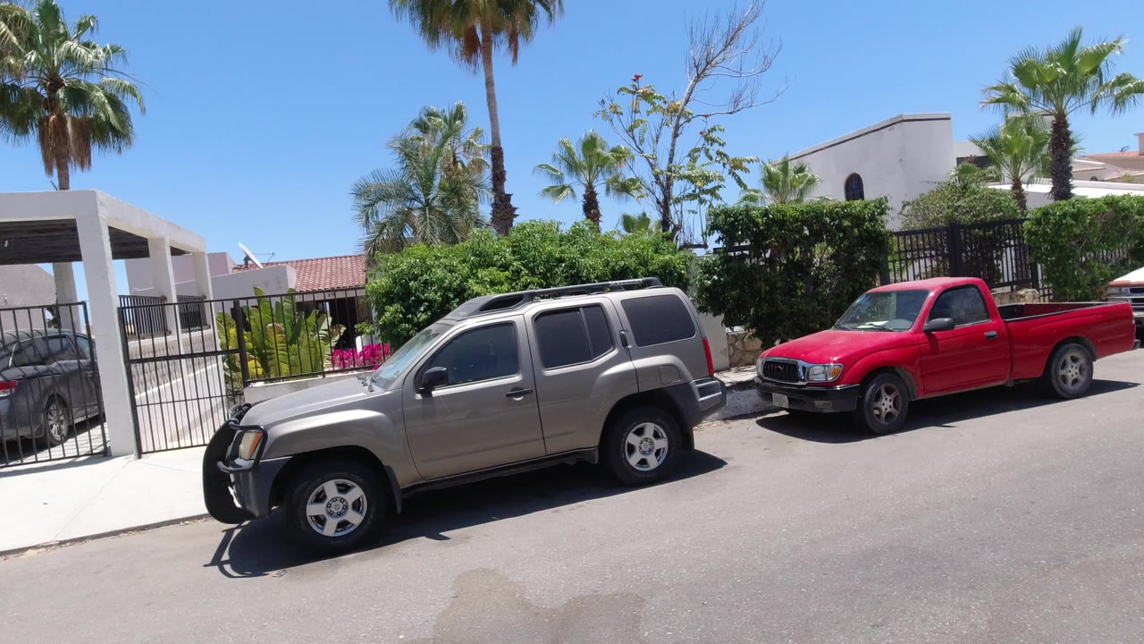 Silver SUV and red pickup parked outside gated white homes under palm trees.