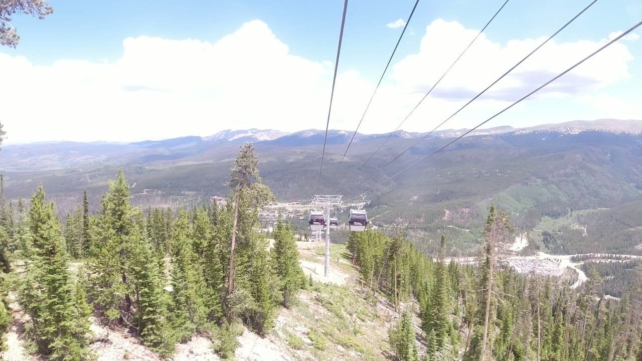 Two gondola cabins travel along cables over a green forested slope in Winter Park, Colorado, with distant ridgelines and no visible people.