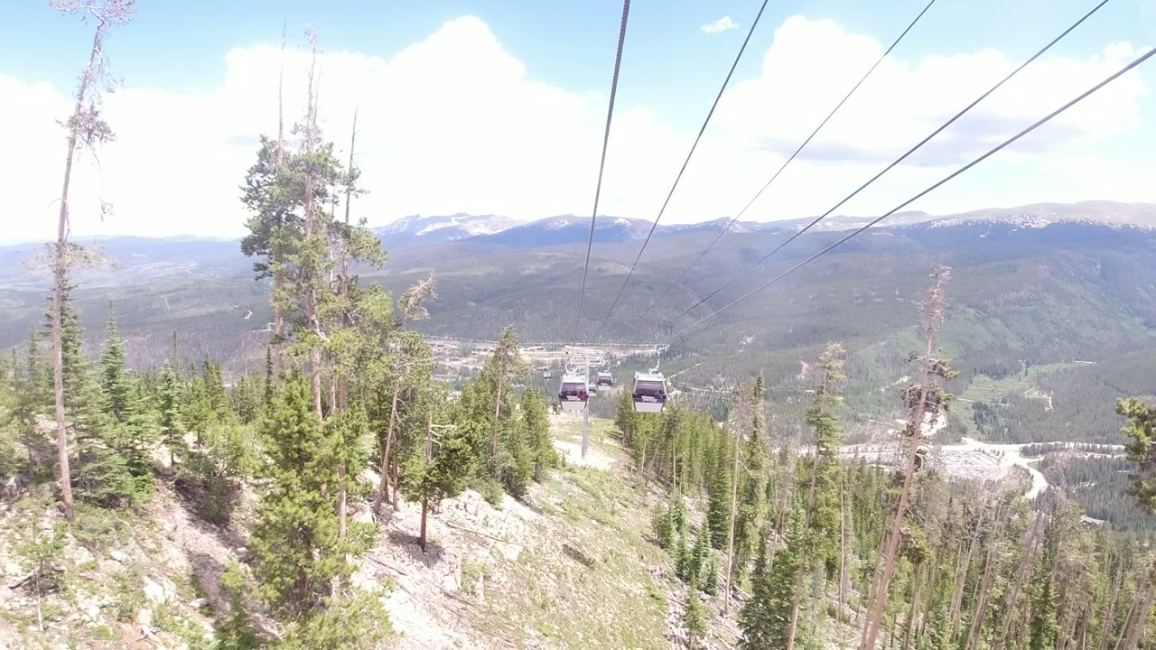 Two gondola cabins appear on the lift line above evergreen trees and a pale rocky hillside in Winter Park, Colorado, with mountains and sky behind them.