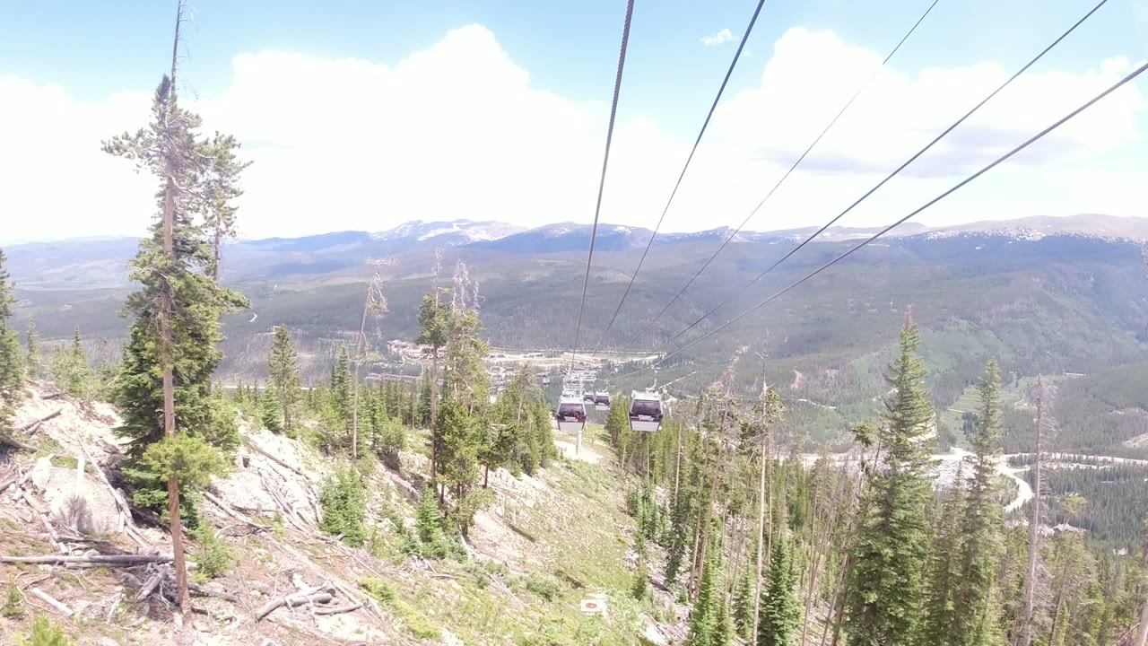 Winter Park, Colorado hillside with tall evergreens, visible gondola cables, small cabins in the distance, hazy mountain ridges, and no visible people.