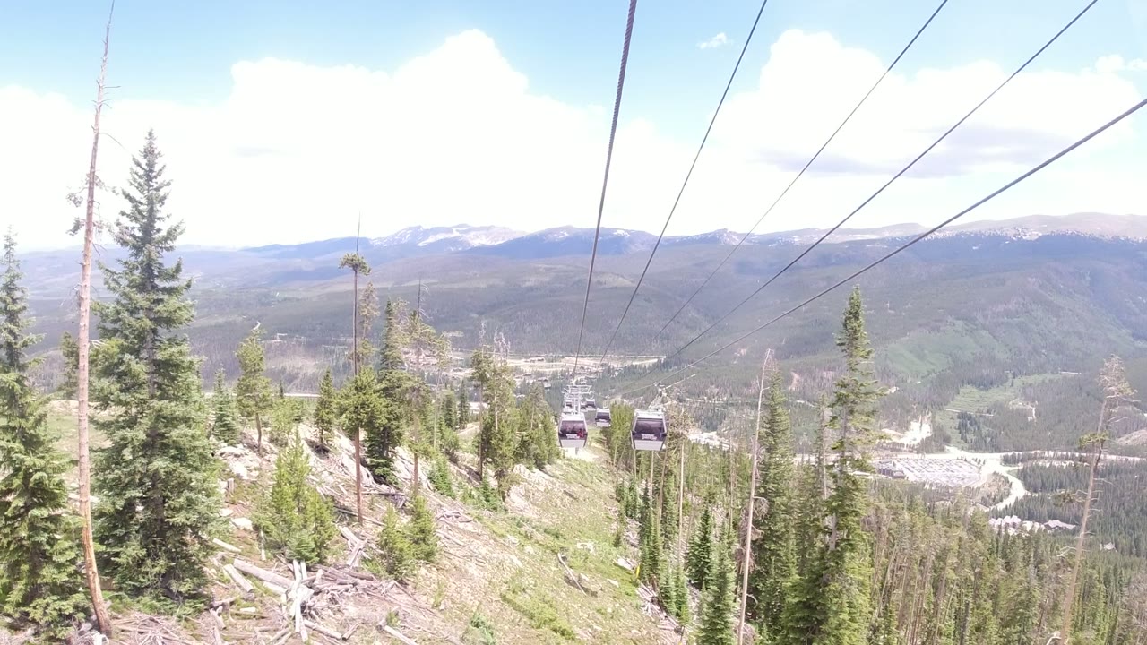 Winter Park, Colorado gondola cabins appear along the cable line above evergreen trees, with pale mountain ridges and a bright sky in the background.
