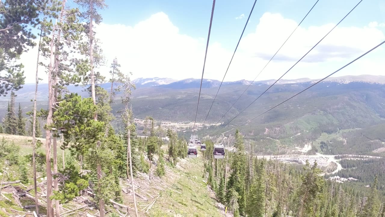 Winter Park, Colorado gondola cabins appear farther down the cable line above pine trees, with a bright sky and layered mountain ridges behind them.