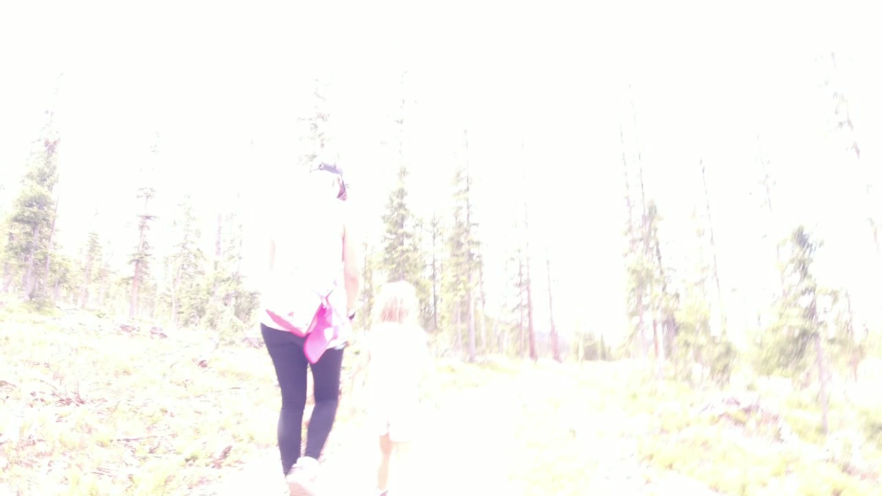 The walkers move farther along the pale trail in Winter Park, Colorado, with a pink garment visible on the adult and the smaller figure washed out by bright light.