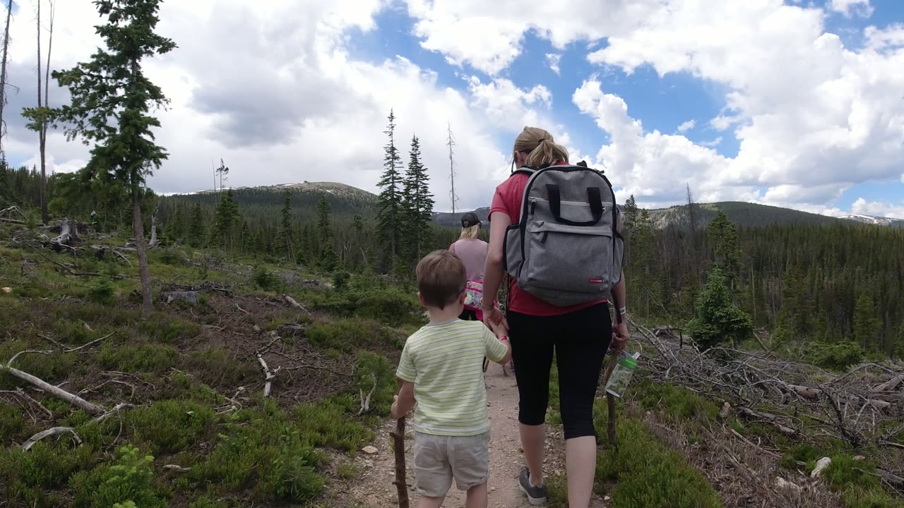 The hikers move farther along the Winter Park trail, with the boy in a pale shirt behind two women and a wide view of pine-covered slopes and broken branches beside the path.