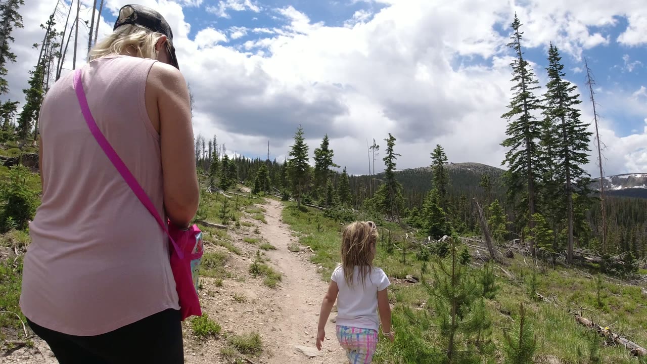 At 0:00 in Winter Park, Colorado, a woman in a pale pink top and a young girl walk away on a dirt path bordered by low pines, dead trunks, rocks, and cloudy sky.