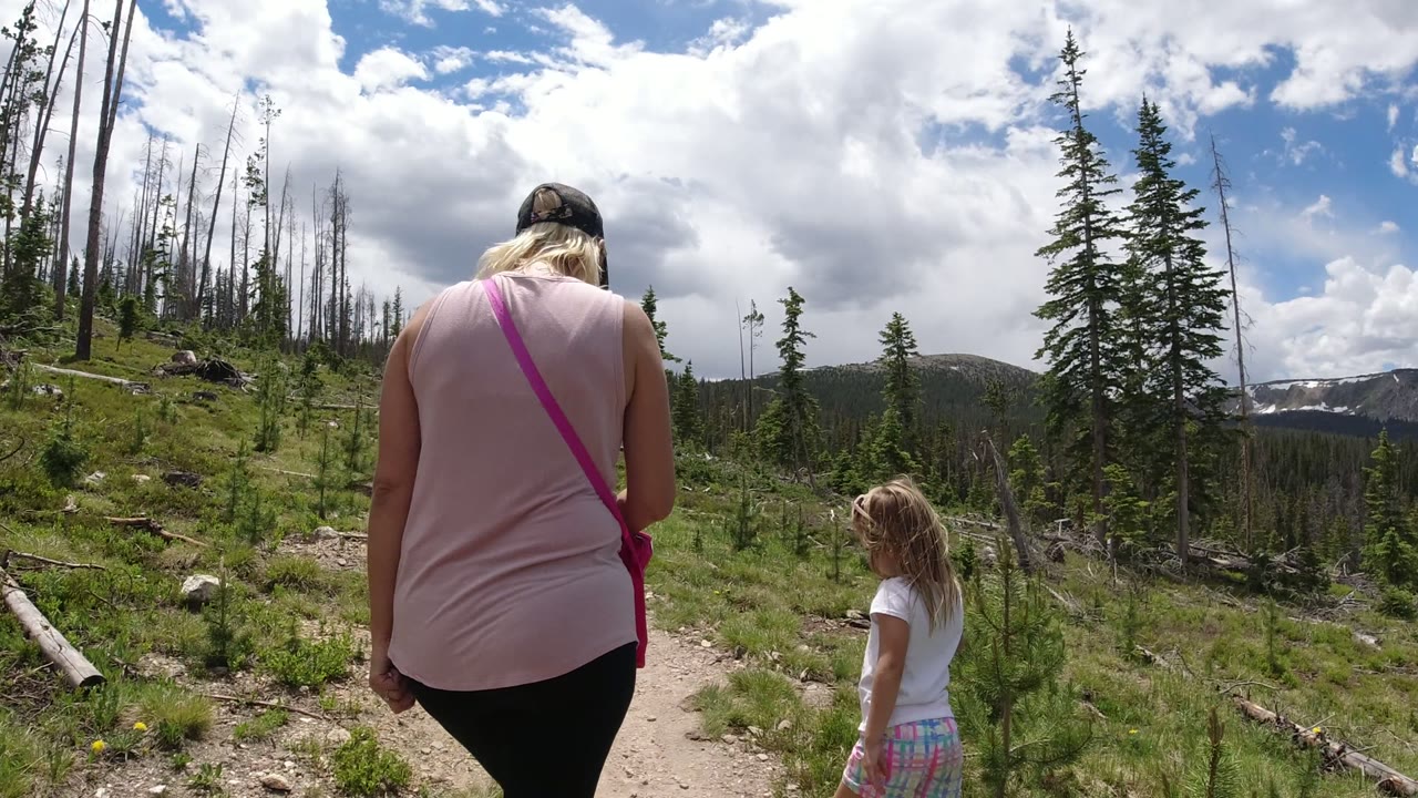 At 0:01 in Winter Park, Colorado, the woman and child are farther up the rocky dirt trail, surrounded by green ground cover, young pines, bare tree trunks, and distant slopes.
