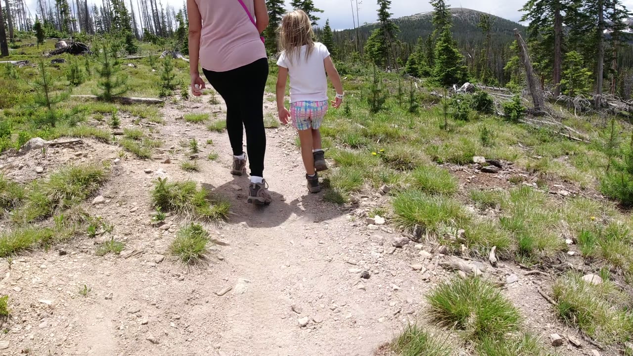 The woman and child are farther along the Winter Park trail, framed from behind with rocky dirt underfoot, scattered grass, pine saplings, and a cloudy bright sky.