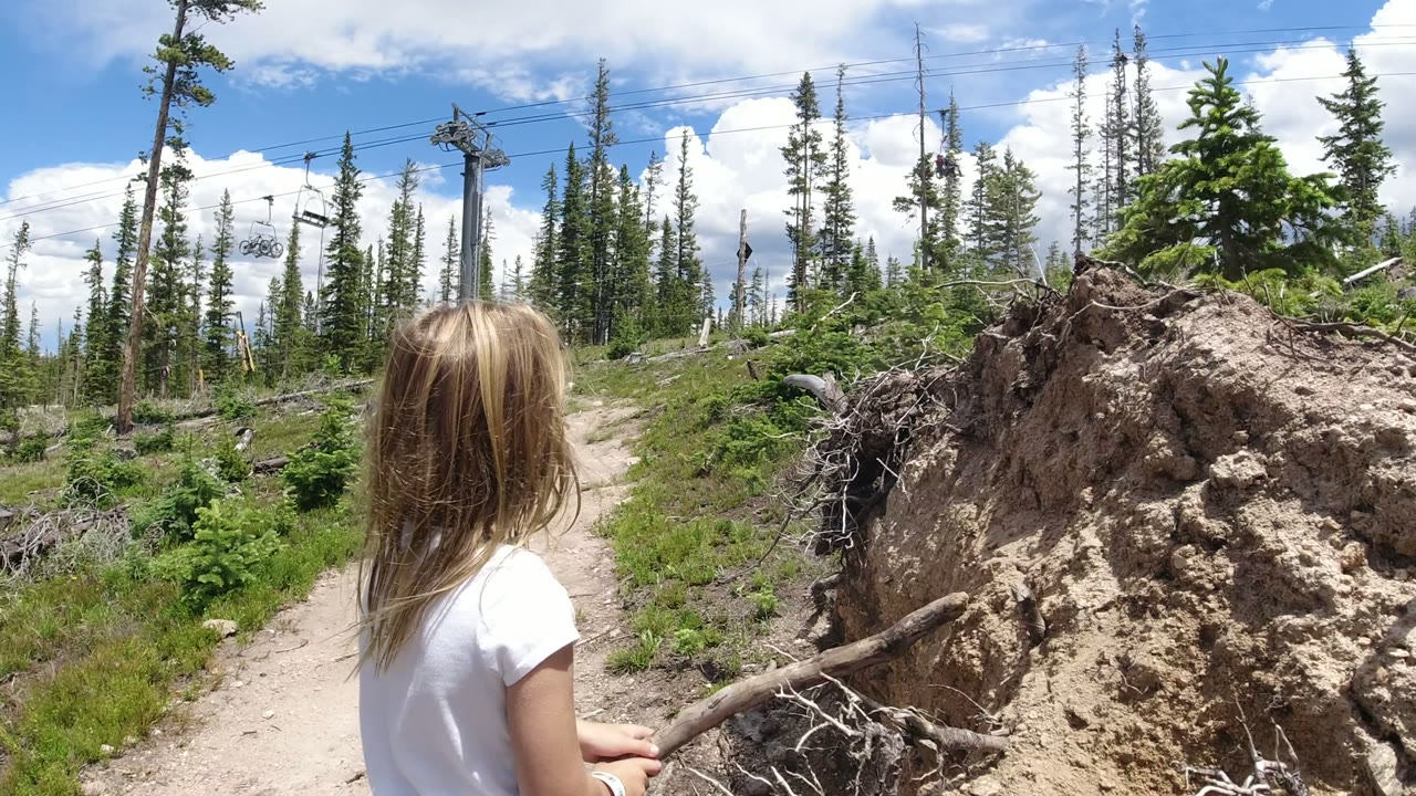 A young blonde girl in a white shirt stands near the left foreground on a Winter Park, Colorado dirt trail, with an uprooted stump, evergreens, lift cables, and bright clouds ahead.