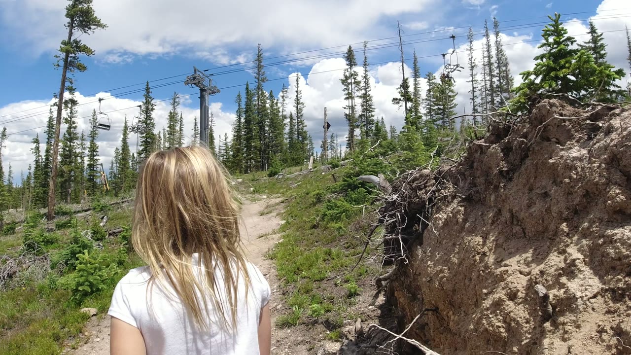 The child is centered from behind on the Winter Park, Colorado trail, with green shrubs along the path, tall evergreens, overhead lift cables, and sunlit clouds in the background.