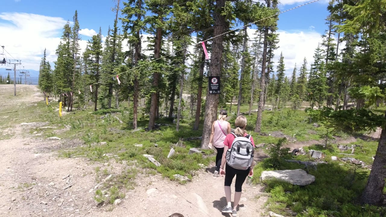 The two hikers continue toward a posted sign on a tree in Winter Park, Colorado, with the dirt path splitting through evergreens and low brush.