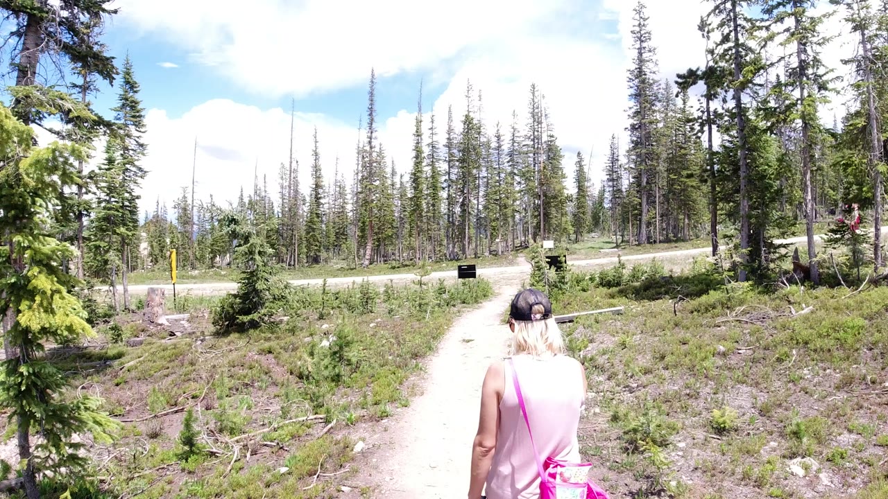 A woman in a pink sleeveless top and black cap walks away from camera on a pale path in Winter Park, Colorado, with conifers and roadside signs ahead.