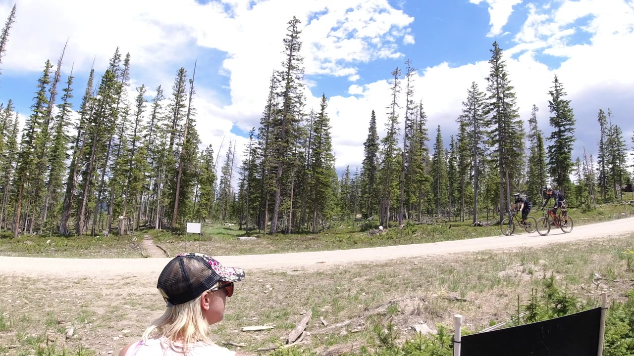 Cyclists ride along a road while a woman in a cap watches from the foreground.