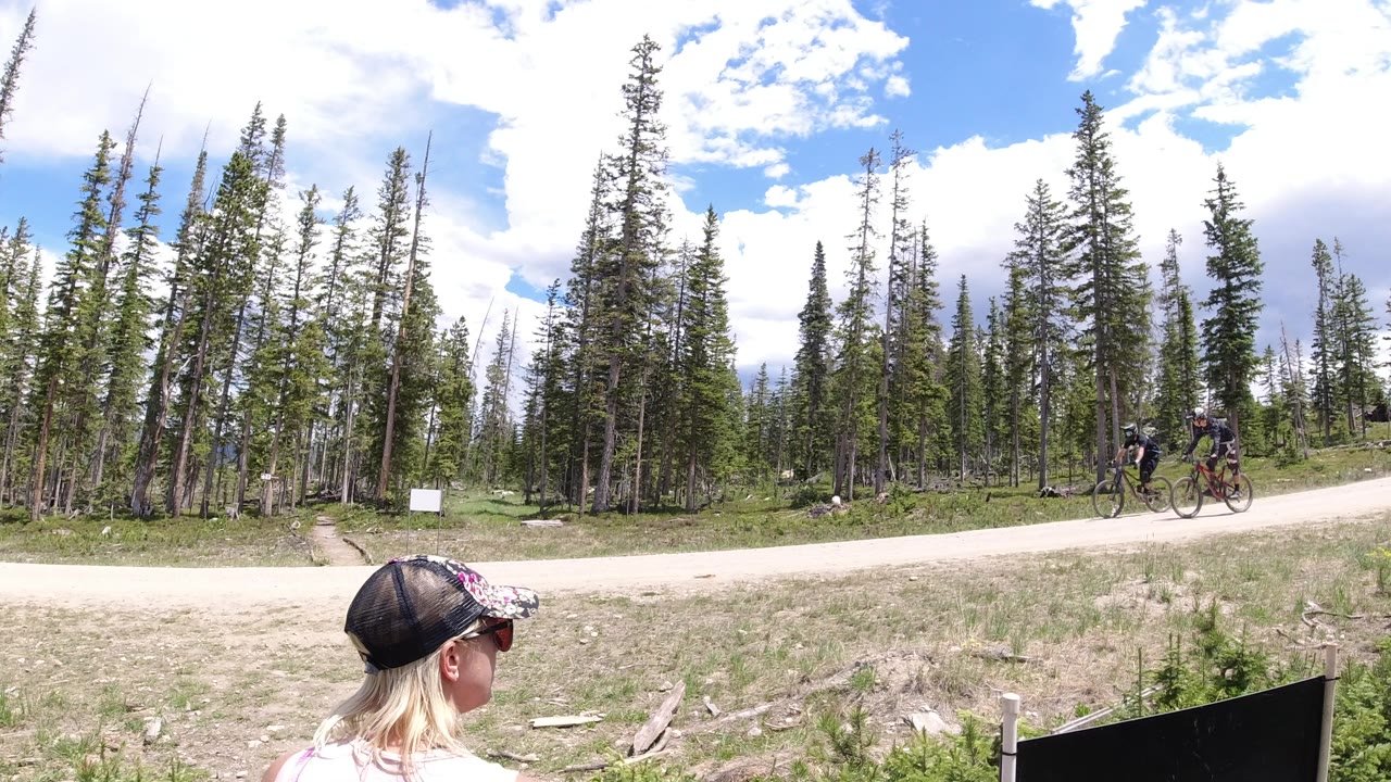 Two riders move along the road in Winter Park, Colorado, with the foreground woman partly visible at the bottom edge and tall pines lining the background.