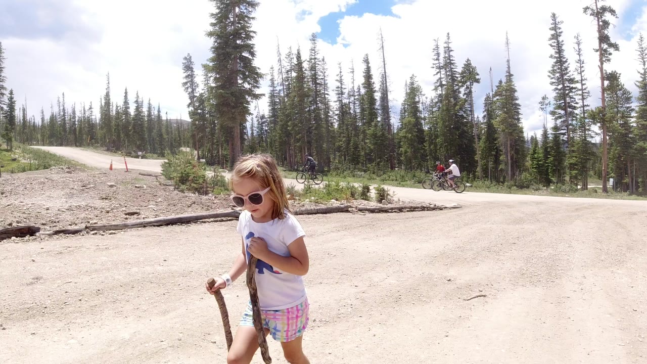 Young girl in sunglasses walks on a dirt path near evergreen trees.