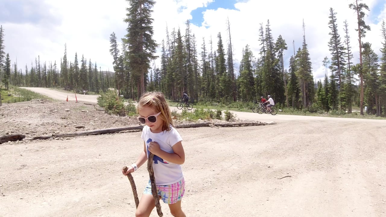 One second in, the girl remains near the lower left foreground on the Winter Park dirt road, with tall trees, a pale road curve, and small cyclists visible in the distance.