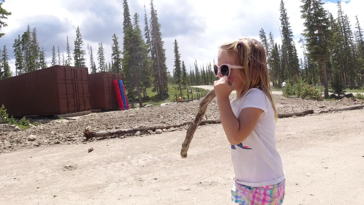 A young girl in sunglasses holds a long stick near her mouth beside a dirt road in Winter Park, Colorado, with tall trees and a red container-like structure behind her.