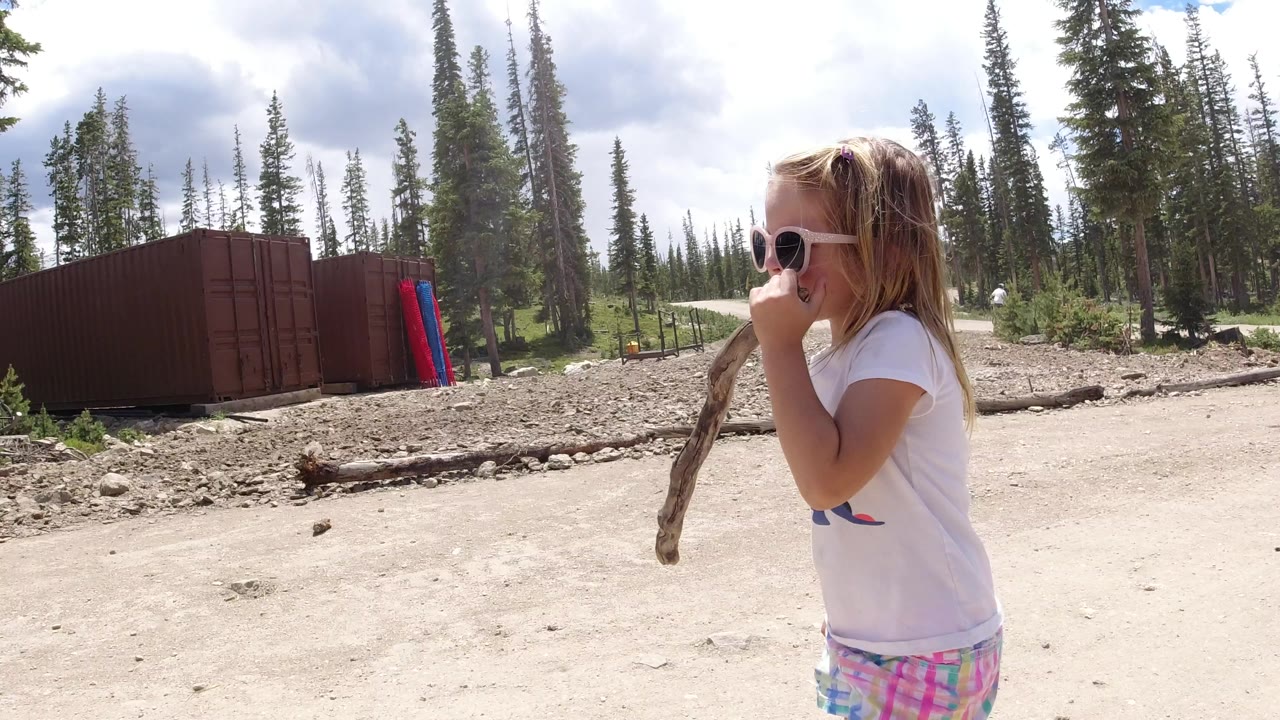 The girl remains near the right side of frame in Winter Park, Colorado, holding the stick while the dirt road curves behind her toward tall trees and a red structure.