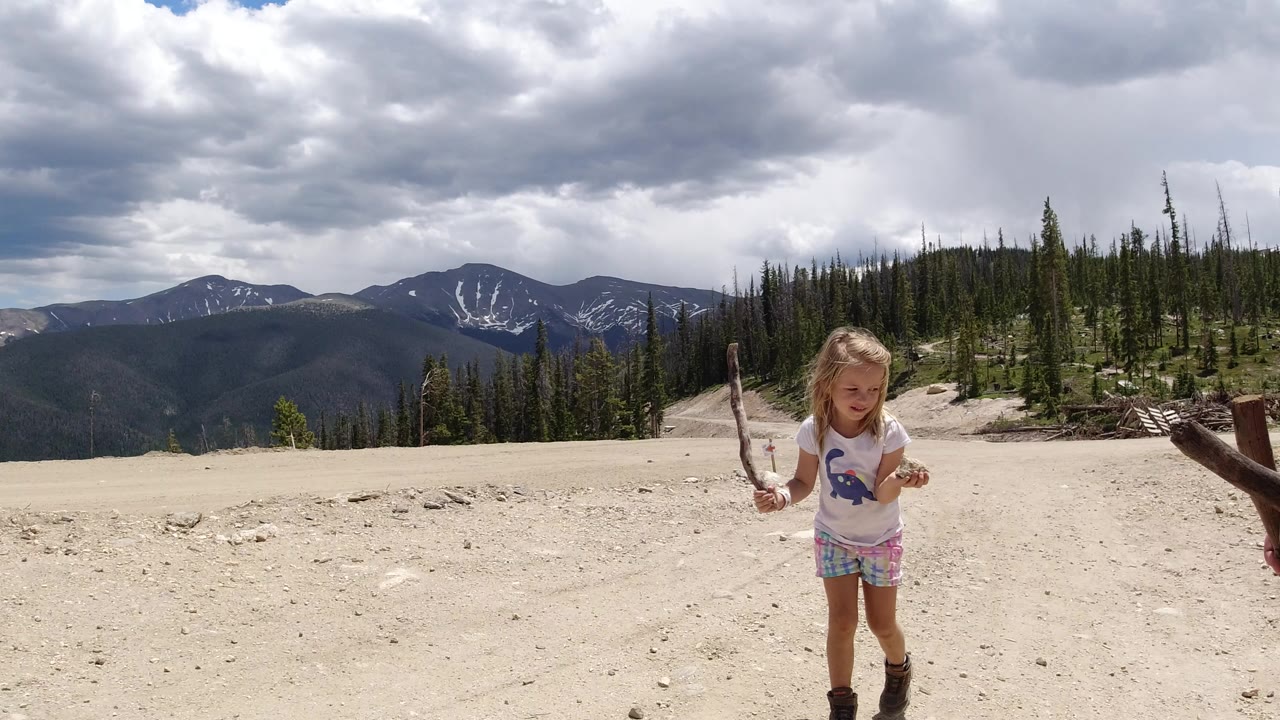 A young girl in Winter Park, Colorado walks across pale sandy terrain holding sticks, with evergreen trees and dark mountain ridges under heavy clouds behind her.