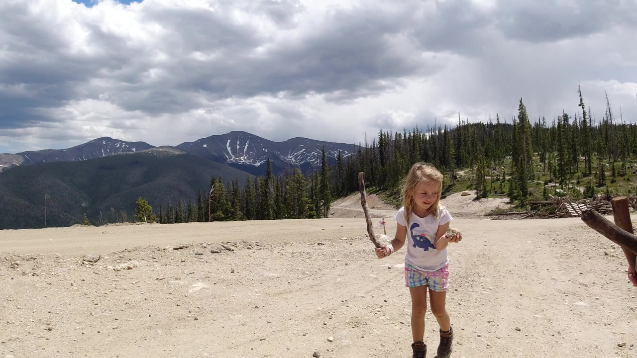 The girl remains near the lower right of the Winter Park, Colorado frame, holding a stick while the sandy foreground opens toward tree-covered slopes and cloud-shadowed mountains.
