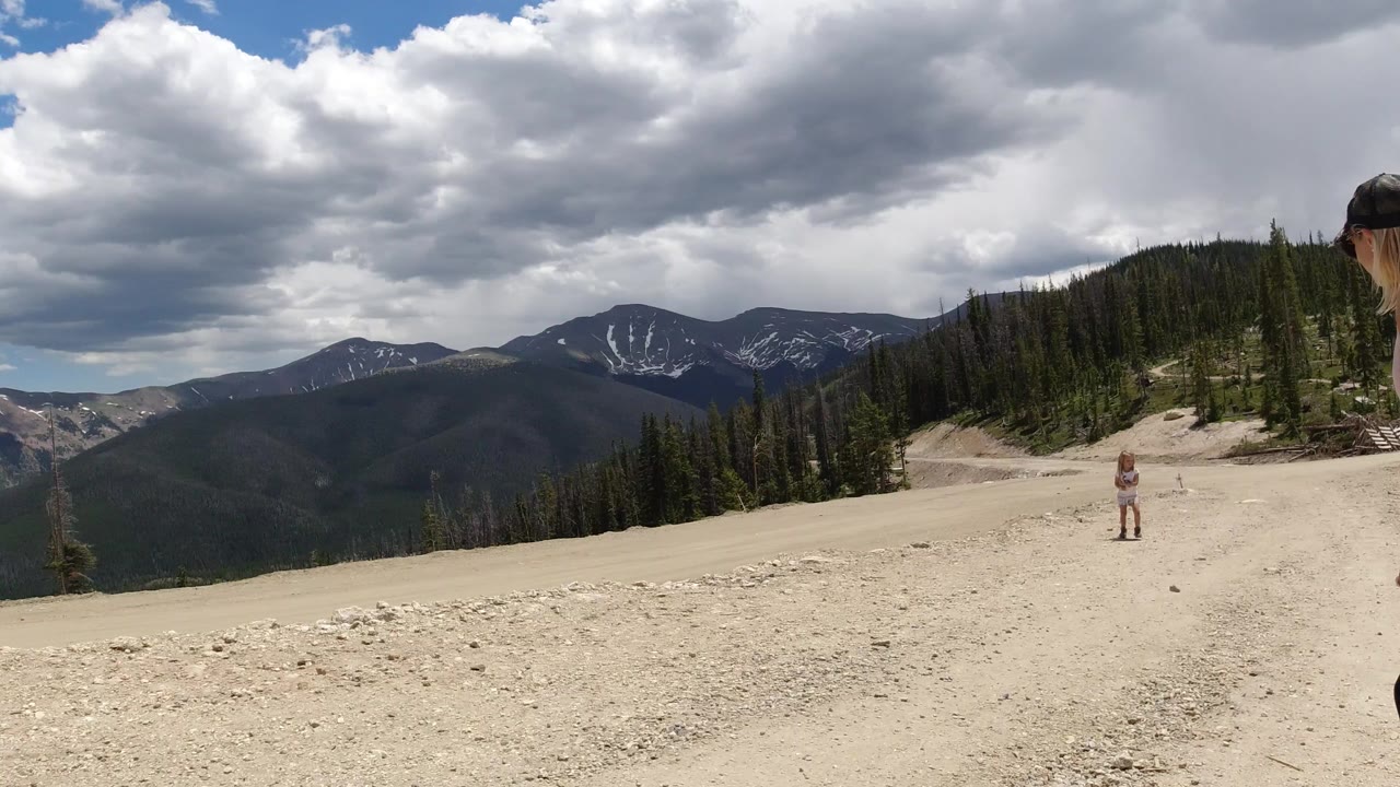 Wide view of a pale rocky road in Winter Park, Colorado, with evergreen slopes, distant snow-streaked mountains, heavy clouds, and no clearly visible people.
