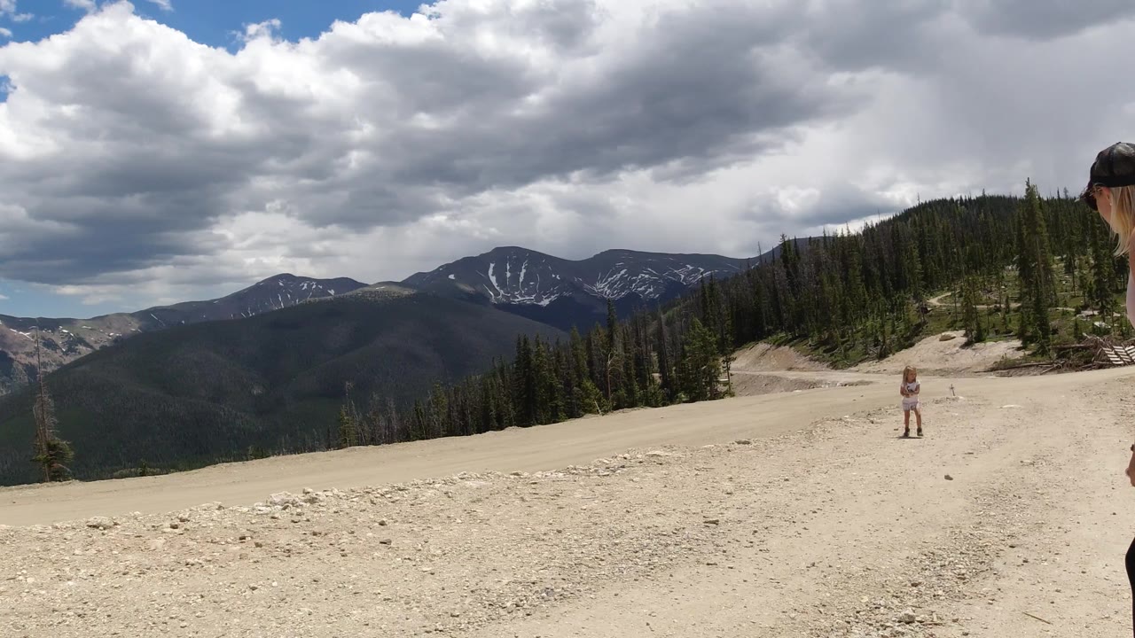 A small hiker stands on the pale rocky road in Winter Park, Colorado, with forested slopes, distant mountains, and large gray clouds overhead.