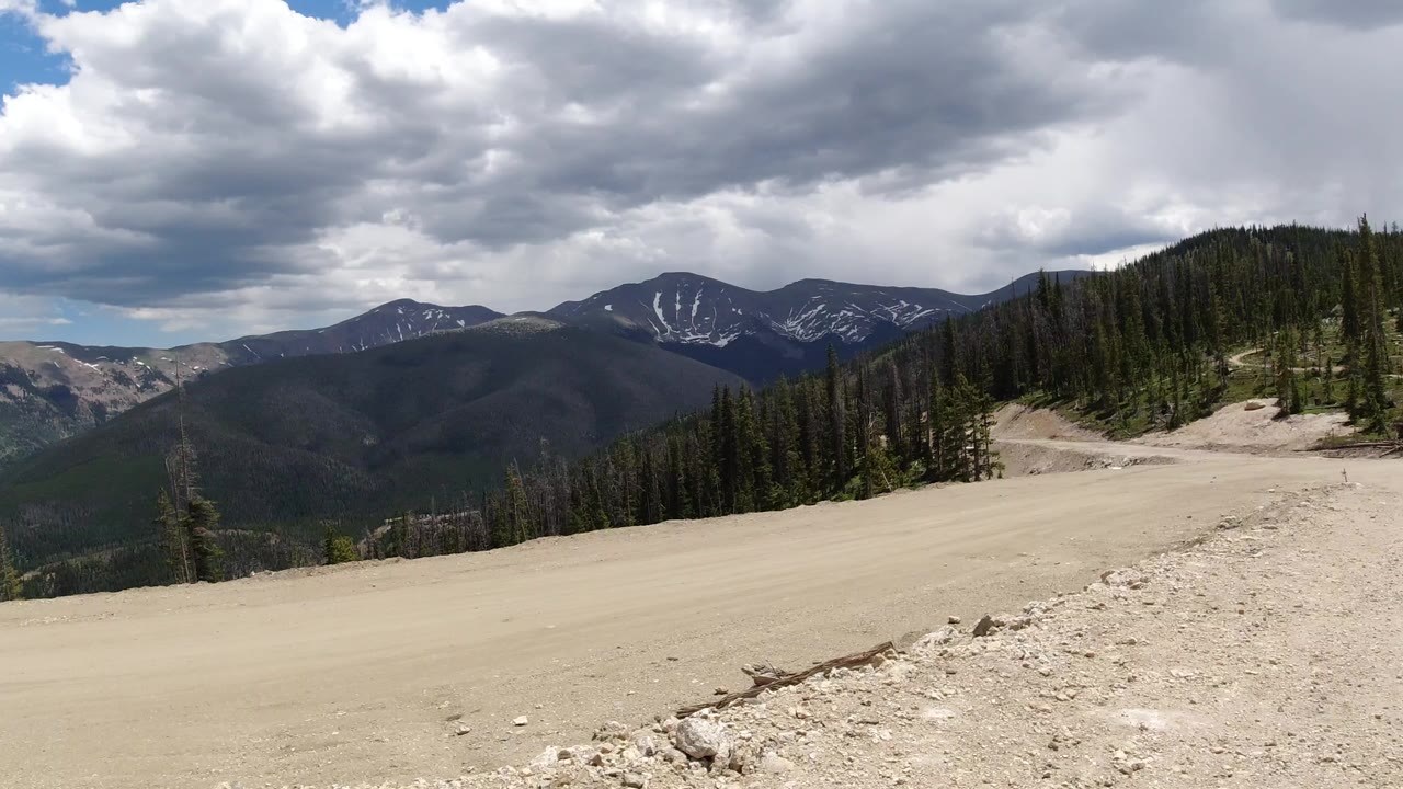 Wide mountain range beyond a pale dirt foreground and evergreen slope.