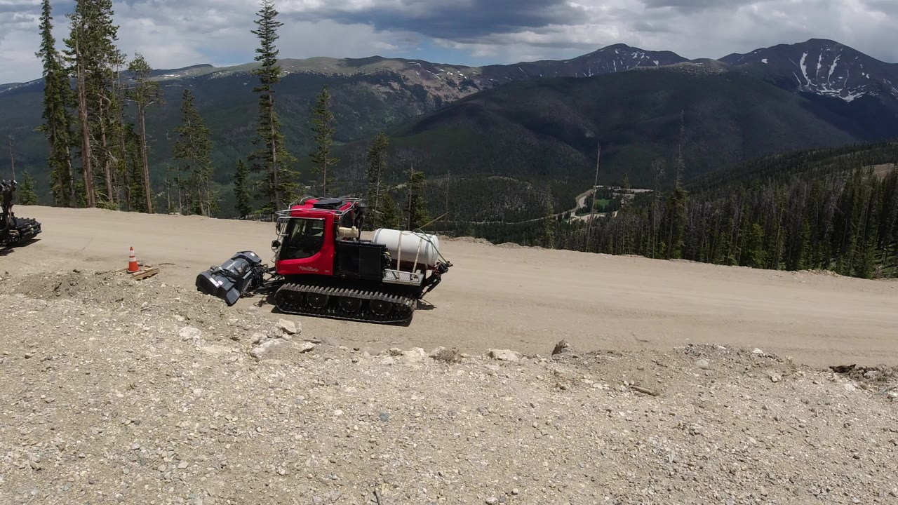 Wide view in Winter Park, Colorado of a red tracked utility vehicle beside a dirt road, rocky foreground, evergreen trees, distant mountain ridges, and no visible people.