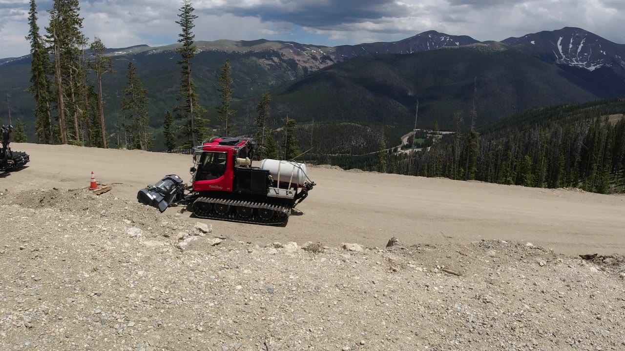 A red tracked work vehicle sits near an orange cone on a pale mountain road in Winter Park, Colorado, with tall evergreens at left and no visible people.