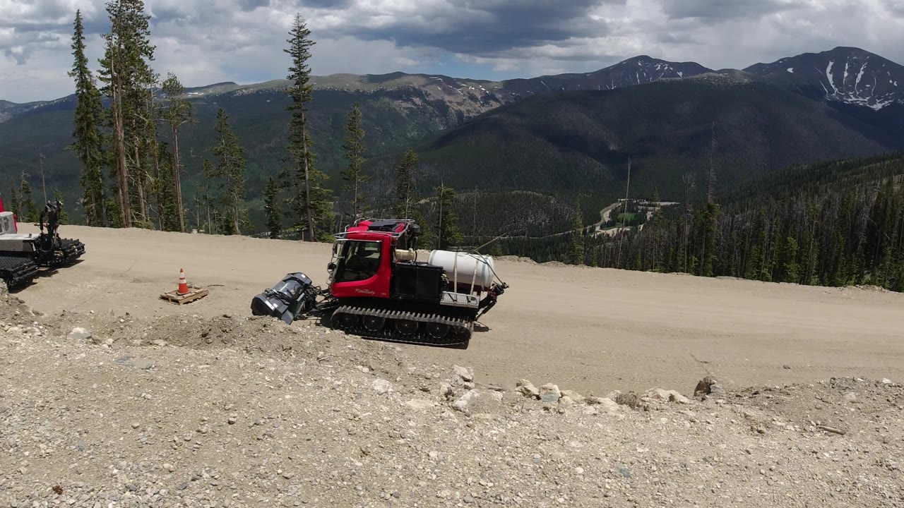 Red mountain service vehicle on a dirt road with evergreen trees and cloudy peaks behind it.