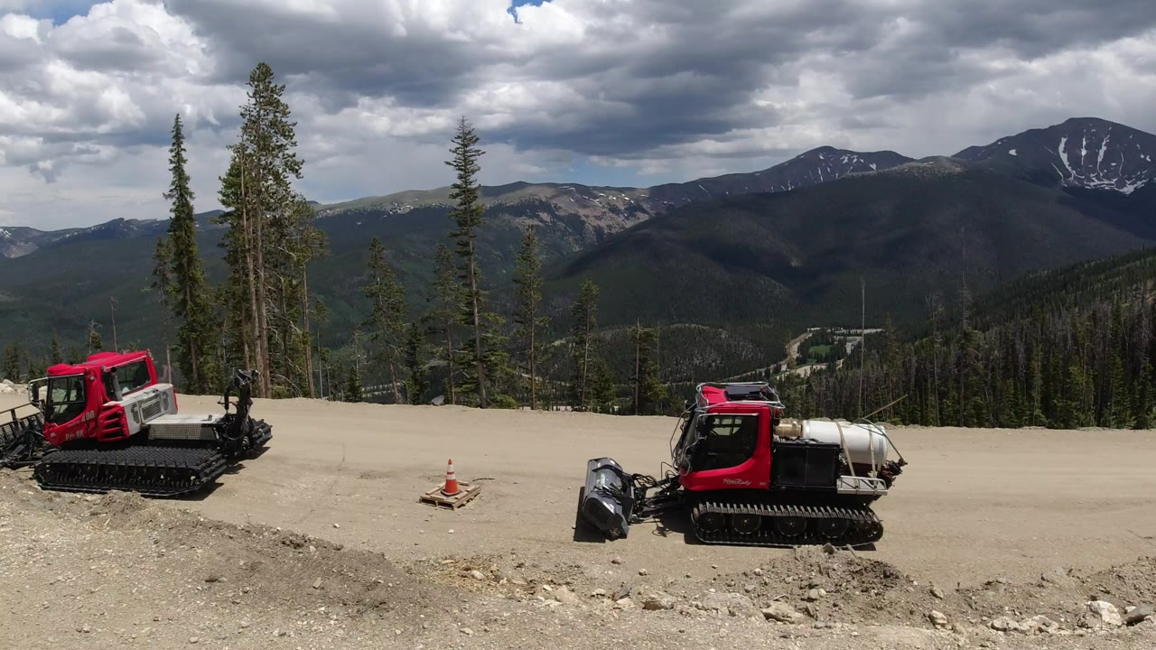 Red tracked construction vehicles sit beside a dirt mountain road in Winter Park, Colorado, with conifers, dark mountain ridges, snow patches, and no visible people.