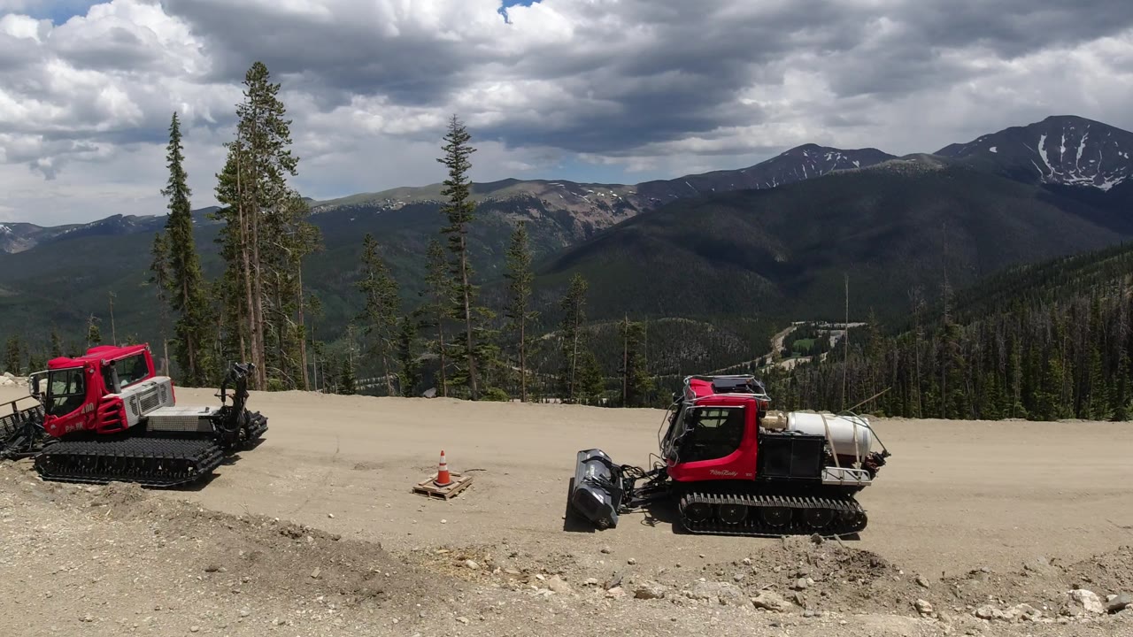 A wide view in Winter Park, Colorado shows two red tracked construction vehicles parked on a pale roadside above forested slopes, with no visible people.