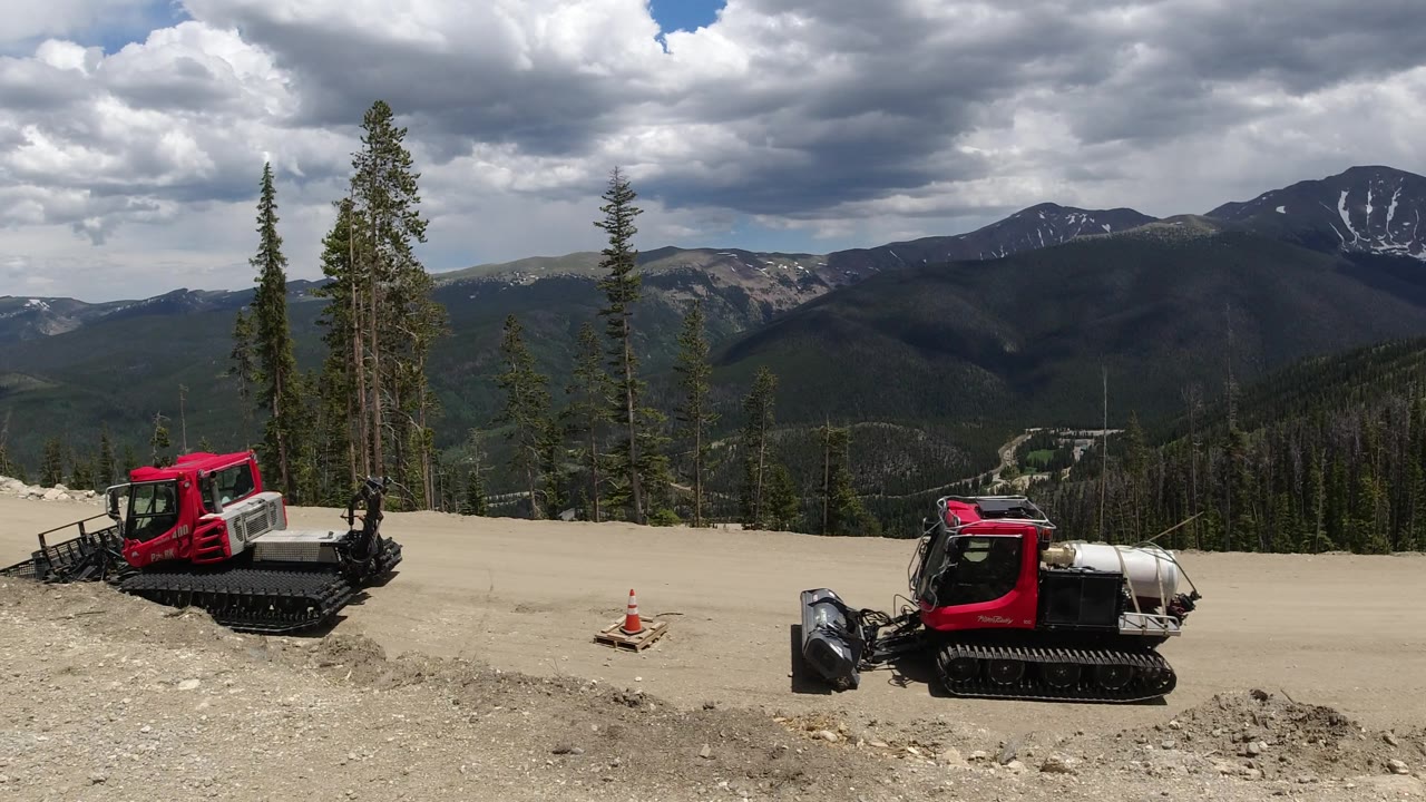 Red tracked construction vehicles, traffic cones, trees, and distant mountain ridges.