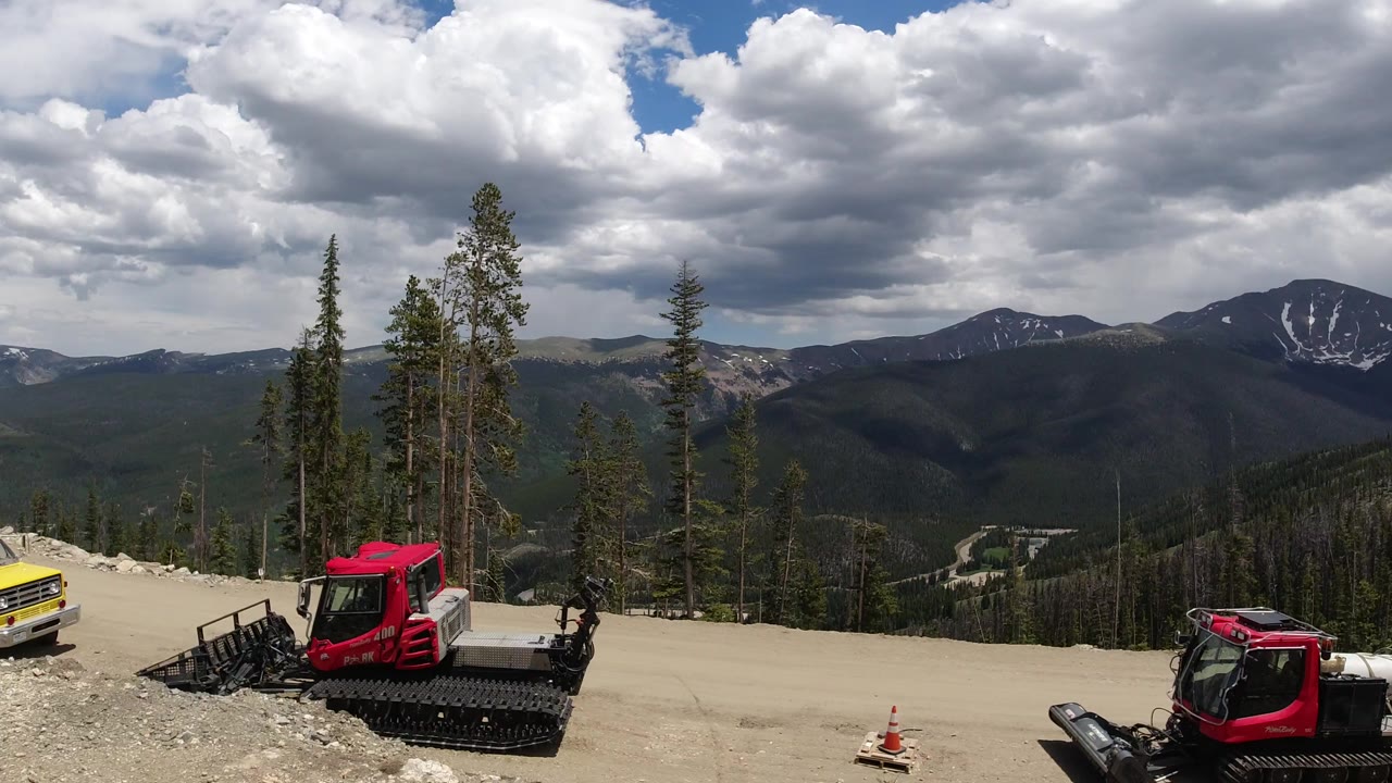 Wide mountain construction area with red tracked equipment and a yellow truck under cloudy sky.